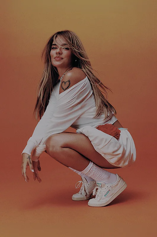 Karol G con outfit blanco y tenis Reebok en campa&ntilde;a sobre fondo naranja