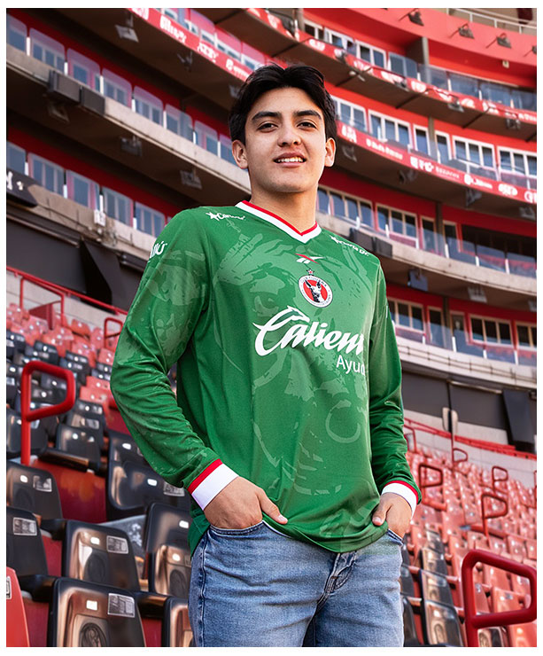 Gilberto Mora posando en gradas del estadio Caliente con jersey verde manga larga Edici&oacute;n Especial Reebok x Club Tijuana, escudo Xoloitzcuintles en pecho, logo Reebok visible, fondo de butacas rojas y negras del estadio, colecci&oacute;n oficial Reebok.