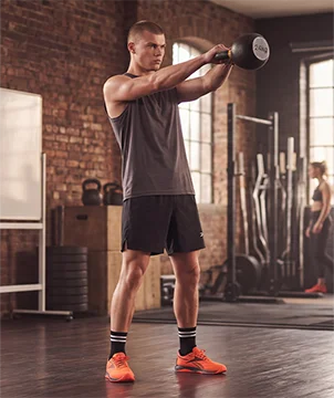 Look mobile de entrenamiento Reebok para hombre, modelo realizando kettlebell swing con pesa rusa de 24 kg en gimnasio de ladrillo, tank top gris, short negro Reebok, calcetines negros con rayas blancas y tenis de training naranjas Reebok Nano, rack de pesas al fondo. Ropa deportiva Reebok para hombre, tenis de training naranjas, outfit de gimnasio y crossfit.