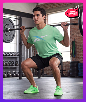 Look Reebok Hot Fashion para hombre, modelo haciendo sentadillas con barra en un gimnasio de ladrillo, playera verde menta Reebok con logo Vector azul, short negro Reebok y tenis de entrenamiento verde ne&oacute;n, kettlebells y discos de peso al fondo, etiqueta Hot Fashion. Reebok hombre, ropa de gimnasio Reebok, playera Reebok, tenis de entrenamiento y rebajas Hot Fashion.