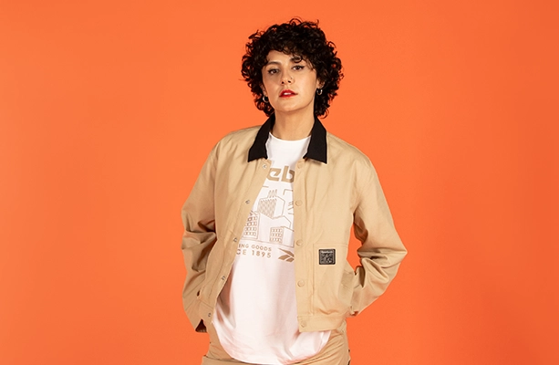 Imagen editorial de la landing Reebok Born Classic Worn Together FW26, modelo sobre fondo naranja con chamarra beige de cuello contrastante y logo Reebok sobre playera blanca con gr&aacute;fico. Reebok, Born Classic Worn Together, ropa Reebok FW26, chamarra Reebok y Reebok Classics