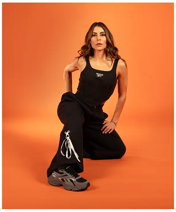 Imagen del carrusel de la landing Reebok Born Classic Worn Together FW26, modelo apoyada en una rodilla sobre fondo naranja con top negro Reebok, pants amplios negros con agujetas y tenis grises. Reebok, Born Classic Worn Together, ropa Reebok FW26, pants Reebok y Reebok Classics.