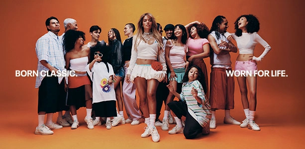 Head banner desktop de landing page Reebok x Karol G Born Classic Worn For Life, grupo diverso de personas de distintas edades y estilos posando en conjunto con Karol G al centro, todos con tenis Reebok Club C 85 blancos sobre fondo naranja. Reebok, Karol G, Club C 85, tenis blancos Reebok, Reebok Classics y campa&ntilde;a Born Classic Worn For Life.