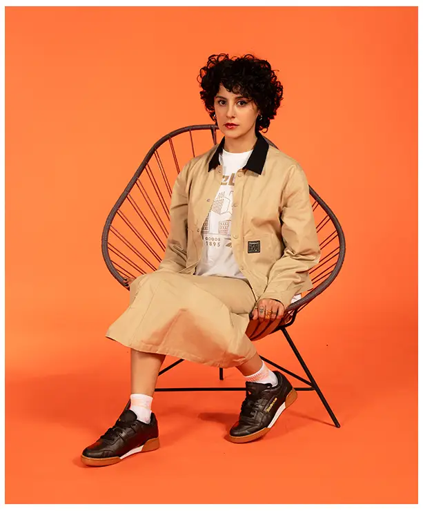 Imagen del carrusel de la landing Reebok Born Classic Worn Together FW26, modelo sentada en una silla Acapulco negra sobre fondo naranja con chamarra beige de cuello contrastante, playera blanca Reebok, falda beige, calcetas blancas y tenis negros. Reebok, Born Classic Worn Together, ropa Reebok FW26, chamarra Reebok y Reebok Classics.