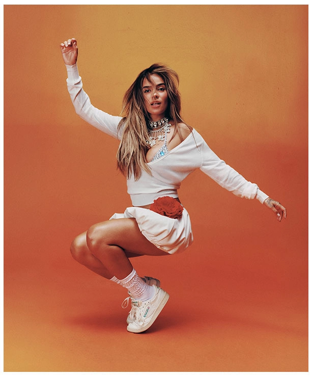 Portrait Reebok x Karol G Born Classic, Karol G bailando con conjunto blanco oversize, collar plateado, calcetines blancos y tenis Reebok Club C 85 blancos sobre fondo naranja. Reebok, Karol G, Club C 85, tenis blancos Reebok, Reebok Classics y campa&ntilde;a Born Classic Worn For Life.