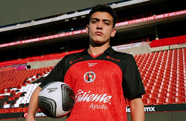 Modelo vistiendo jersey Reebok de Club Tijuana rojo y negro con bal&oacute;n en el estadio