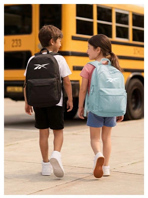 Portrait Reebok Back to School, ni&ntilde;o con mochila negra Reebok y ni&ntilde;a con mochila azul claro Reebok de espaldas caminando hacia autob&uacute;s escolar amarillo, ambos con tenis blancos. Mochilas Reebok, tenis blancos para ni&ntilde;os, tenis para la escuela y regreso a clases
