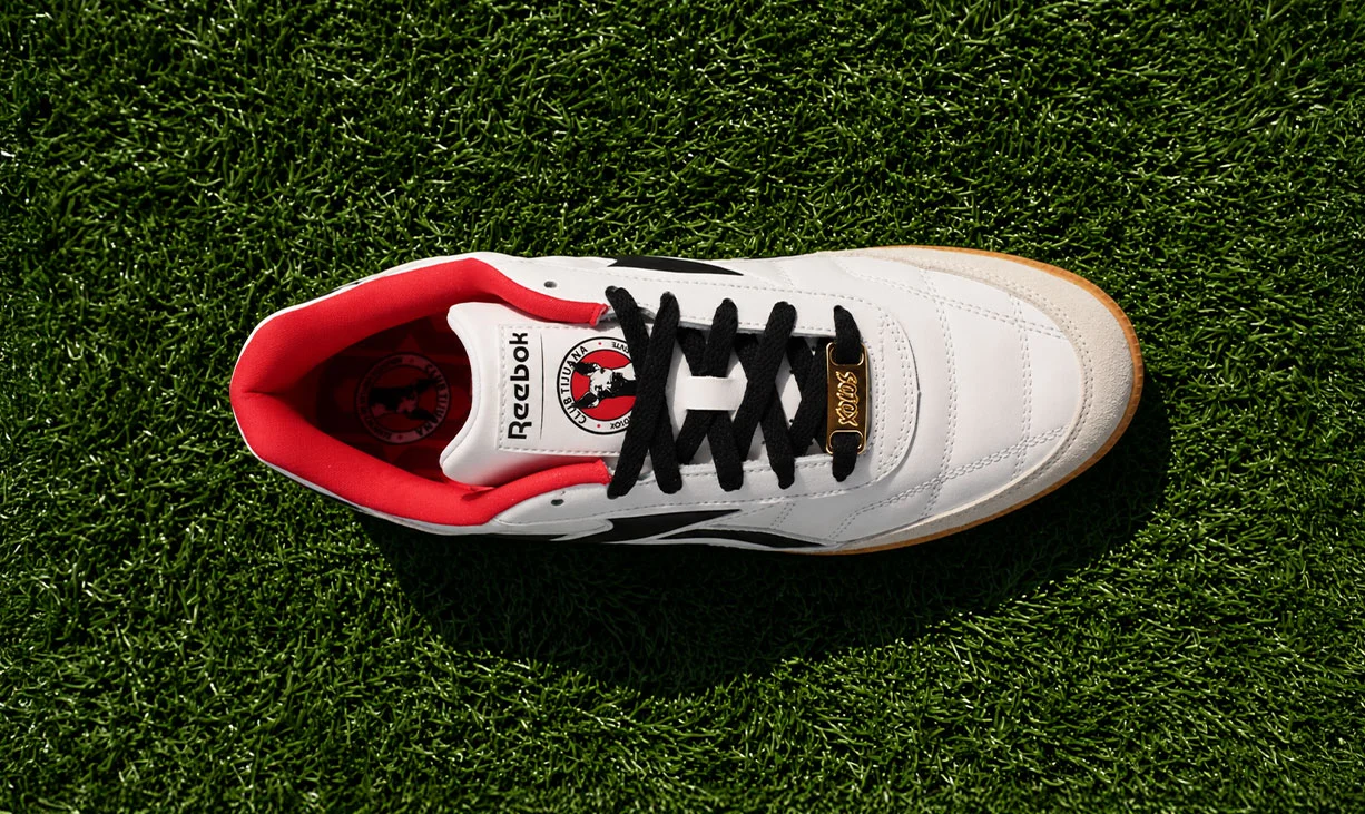 Vista superior de un tenis Reebok Finale x Xolos blanco de piel con agujetas negras, forro rojo, logo Reebok Vector negro, herraje dorado Xolos y escudo del Club Tijuana en la lengua, sobre pasto verde. Reebok, Xolos, Club Tijuana, tenis Reebok Finale, tenis retro de f&uacute;tbol y colaboraci&oacute;n Reebok x Xolos.