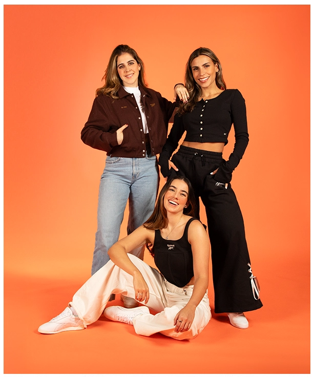 Imagen del carrusel de la landing Reebok Born Classic Worn Together FW26, tres amigas sobre fondo naranja, una con chamarra vino y jeans, otra con top negro y pants negros Reebok y una tercera sentada con top negro y pants color crema. Reebok, Born Classic Worn Together, ropa Reebok FW26, looks Reebok y Reebok Classics